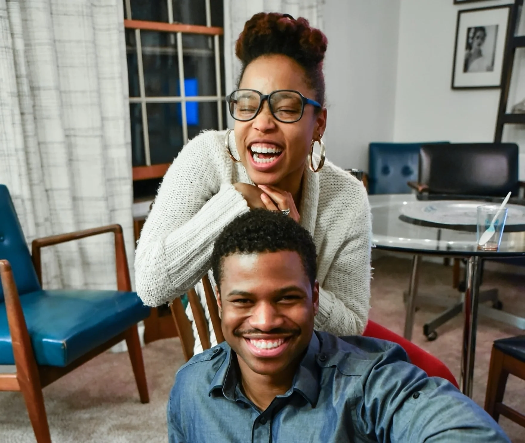 Two people laughing together in a living room: a woman in glasses and a cream knit sweater leans over the shoulders of a smiling man in a denim shirt.